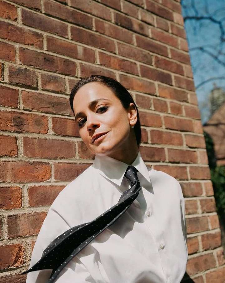 Woman in a white shirt and polka dot tie, posing in front of a brick wall in natural light, creating a sophisticated and modern look.