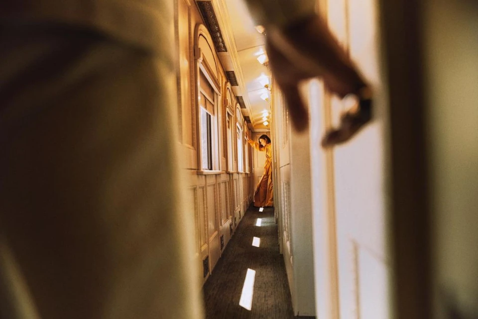 Woman in an ochre gown stands in the aisle of a vintage train car as a partial hand reaches toward her in the foreground.