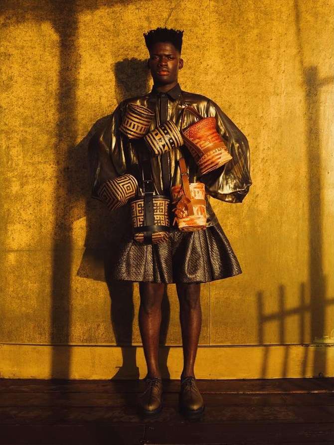 Stylish man poses in front of textured yellow wall, wearing metallic coat, pleated skirt, and an array of woven patterned bags.