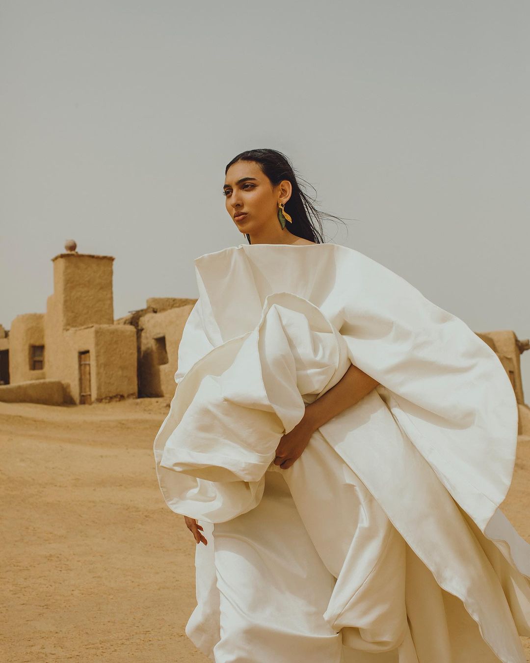 An adult female wearing a white dress stands in the desert in front of buildings in a wide shot captured in an outdoor setting.