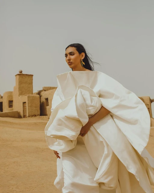 An adult female wearing a white dress stands in the desert in front of buildings in a wide shot captured in an outdoor setting.