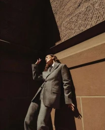 Low-angle shot of a woman in a grey wool suit posing against a warm stone building under harsh sunlight, photographed by Gleeson Paulino.