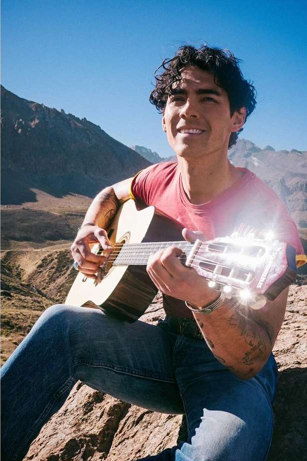 A man with dark curly hair sitting outdoors playing an acoustic guitar against a mountainous backdrop in bright sunlight