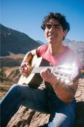 A man with dark curly hair sitting outdoors playing an acoustic guitar against a mountainous backdrop in bright sunlight