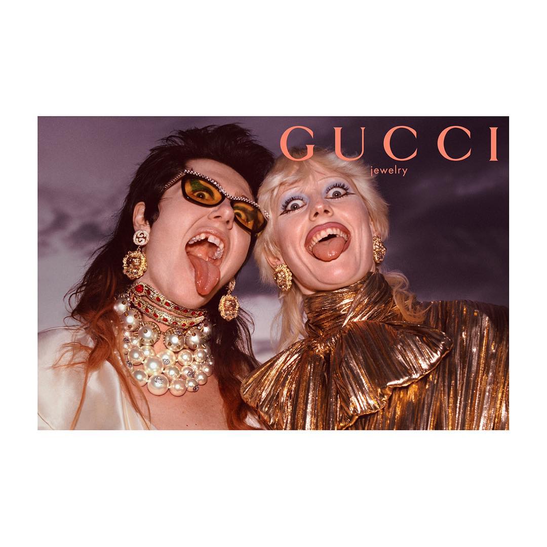 Two women with tongues out wearing Gucci jewelry, gold tops, and statement earrings against a soft background, bold brand name.