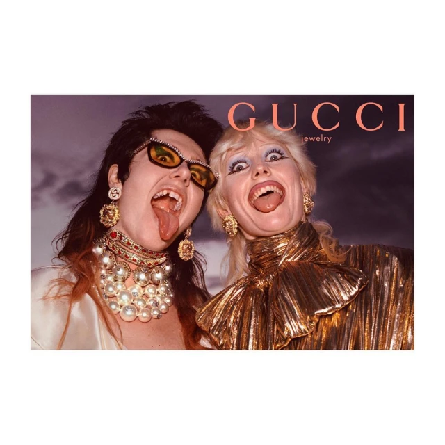 Two women with tongues out wearing Gucci jewelry, gold tops, and statement earrings against a soft background, bold brand name.