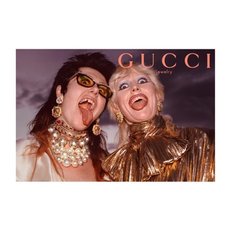 Two women with tongues out wearing Gucci jewelry, gold tops, and statement earrings against a soft background, bold brand name.