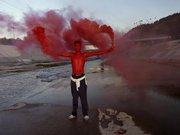 Adult stands in a waterway with red smoke flares. The setting is outdoors with some hillside and an underpass in the background.
