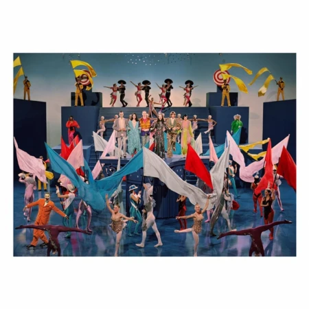 Stage performance featuring group of dancers and actors on tiered platform with colorful costumes, flags, and theatrical lighting in a theatre.