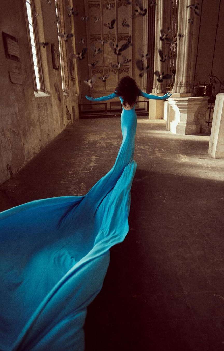 Woman in turquoise gown walks toward light, arms outstretched, surrounded by birds in aged cathedral. Fashion, beauty, and fine art.