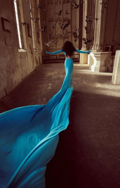 Woman in turquoise gown walks toward light, arms outstretched, surrounded by birds in aged cathedral. Fashion, beauty, and fine art.