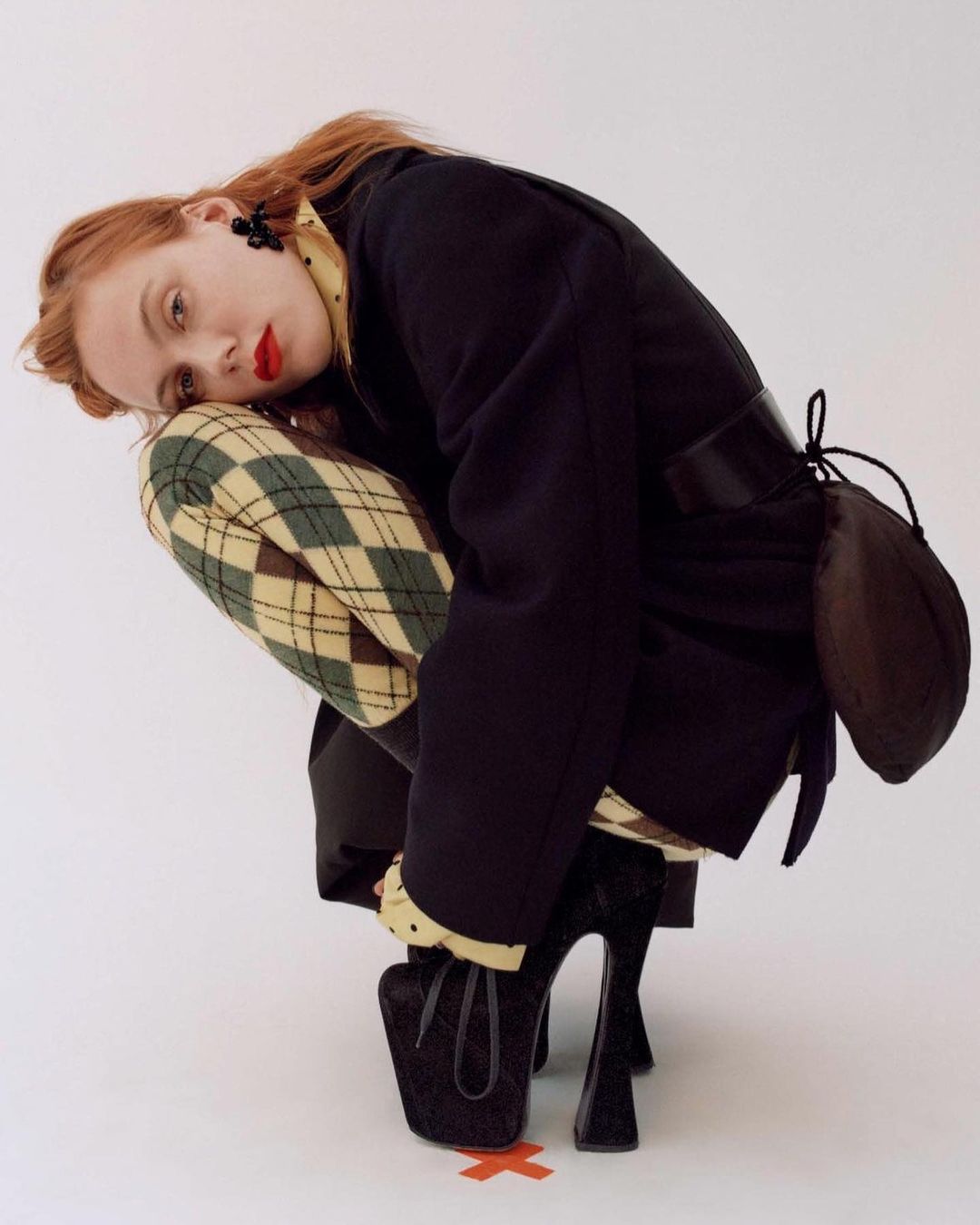 Fashion portrait of a woman with red hair crouching in a black jacket, patterned shirt, and patterned trousers, with black platform heels.