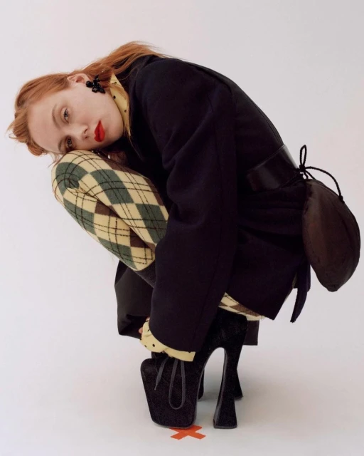 Fashion portrait of a woman with red hair crouching in a black jacket, patterned shirt, and patterned trousers, with black platform heels.