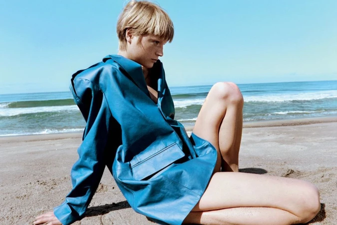 Model wearing a blue leather jacket sitting on a sandy beach by the ocean under a clear sky.