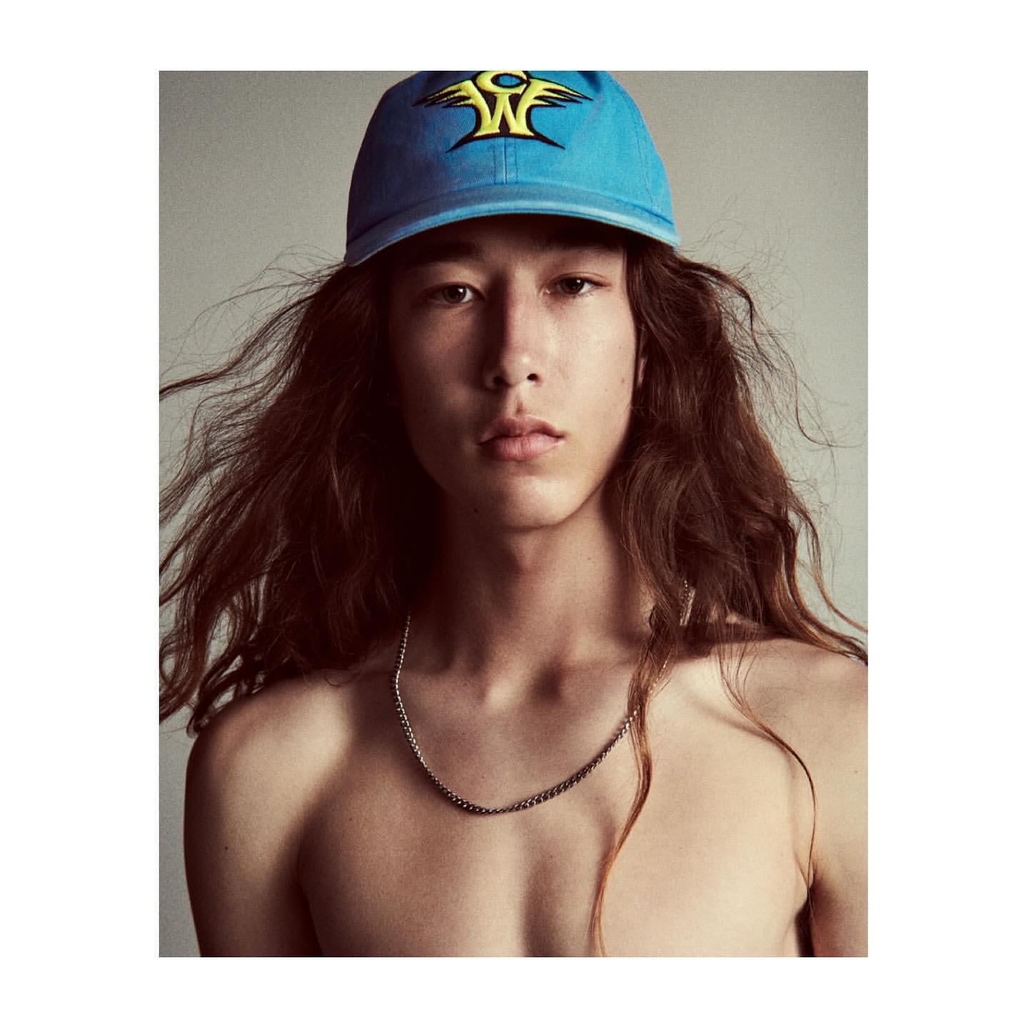 Portrait of a young adult man with long brown hair wearing a blue baseball cap with a yellow design, posing in a photo studio.