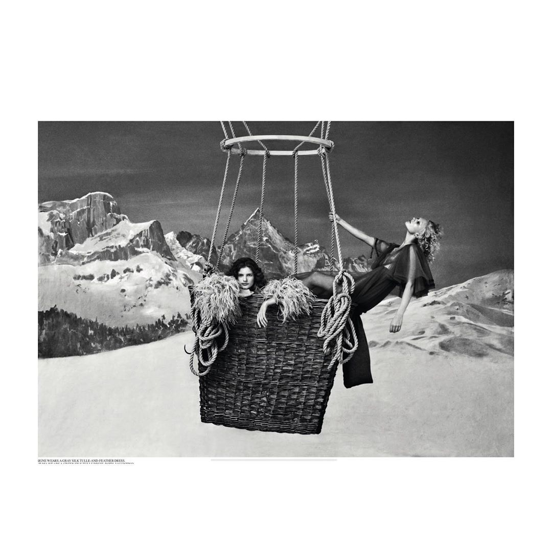 Two women in sheer outfits pose inside of a hot air balloon basket with rope against a backdrop of snowy mountains in black and white.