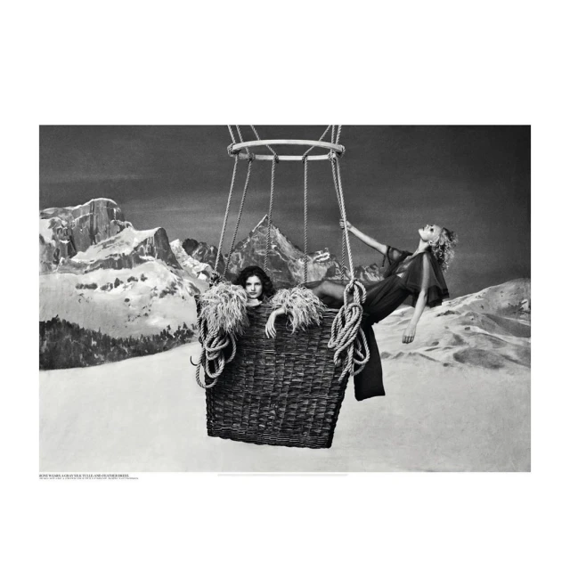 Two women in sheer outfits pose inside of a hot air balloon basket with rope against a backdrop of snowy mountains in black and white.