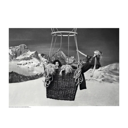 Two women in sheer outfits pose inside of a hot air balloon basket with rope against a backdrop of snowy mountains in black and white.