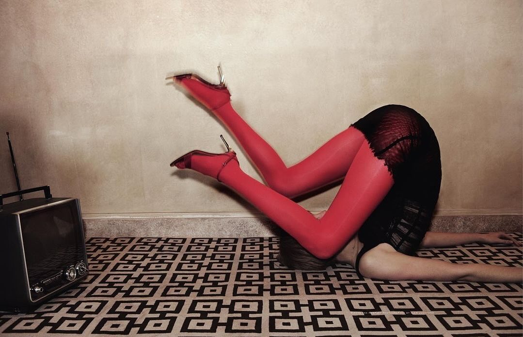 A woman in red tights and high heels bends over backwards on a patterned rug, with a vintage-style television set to the side.