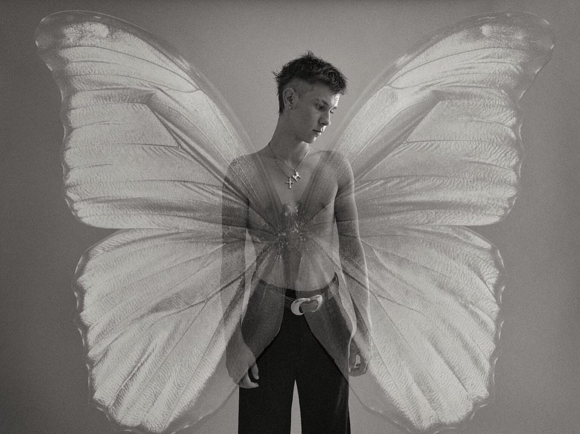 Portrait of a shirtless adult male with a cross necklace, looking down, set against large superimposed butterfly wings in a studio setting.