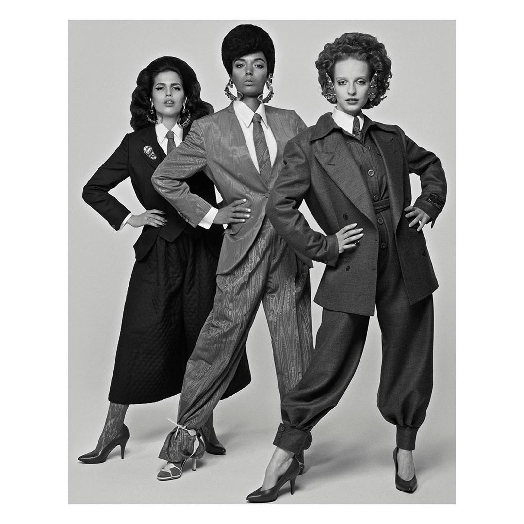 Three women in suits stand in a studio with neutral lighting. The black and white photo features models in vintage-inspired power suits.