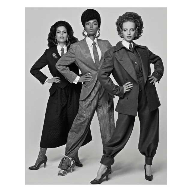 Three women in suits stand in a studio with neutral lighting. The black and white photo features models in vintage-inspired power suits.