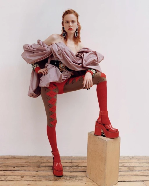 A redhead fashion model poses indoors wearing platform shoes, patterned tights, and a mauve ruffled top against a white background.