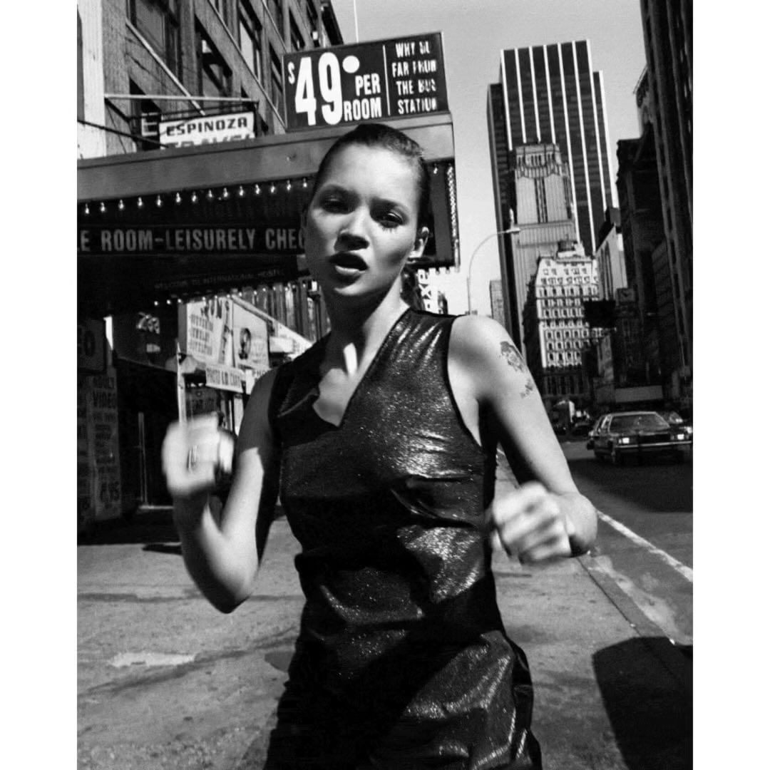 Black and white portrait of Kate Moss in a dress, standing in front of a building, captured at eye-level in an urban environment.
