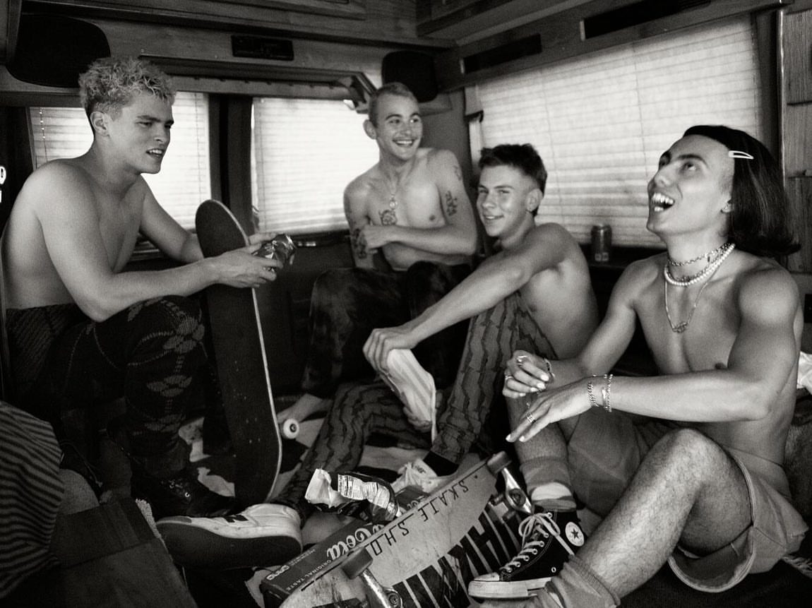 Black and white photograph of four shirtless young men sitting inside a vehicle with skateboards and footwear scattered around them.