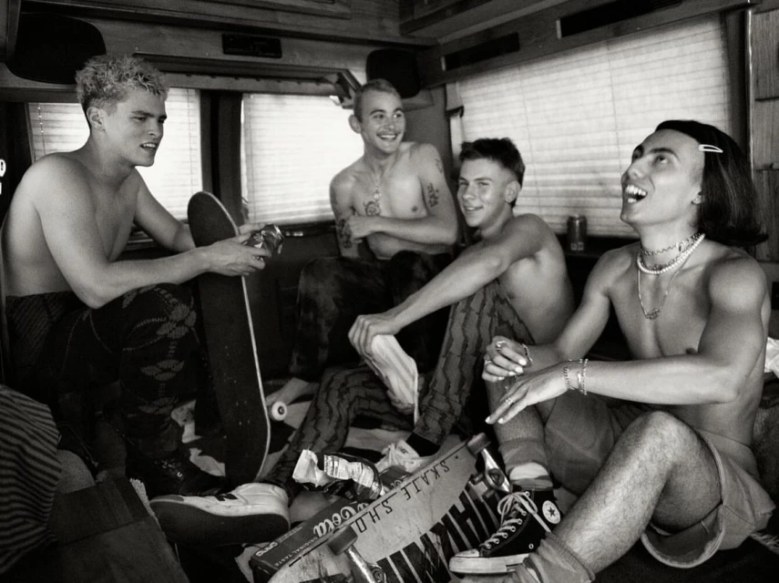 Black and white photograph of four shirtless young men sitting inside a vehicle with skateboards and footwear scattered around them.
