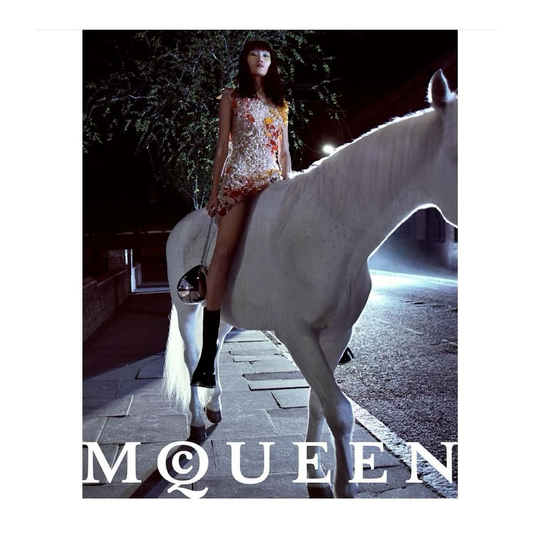 Fashion shot of a slender Asian woman in a dress, riding a white horse at night, under bright light, with 'McQueen' logo.