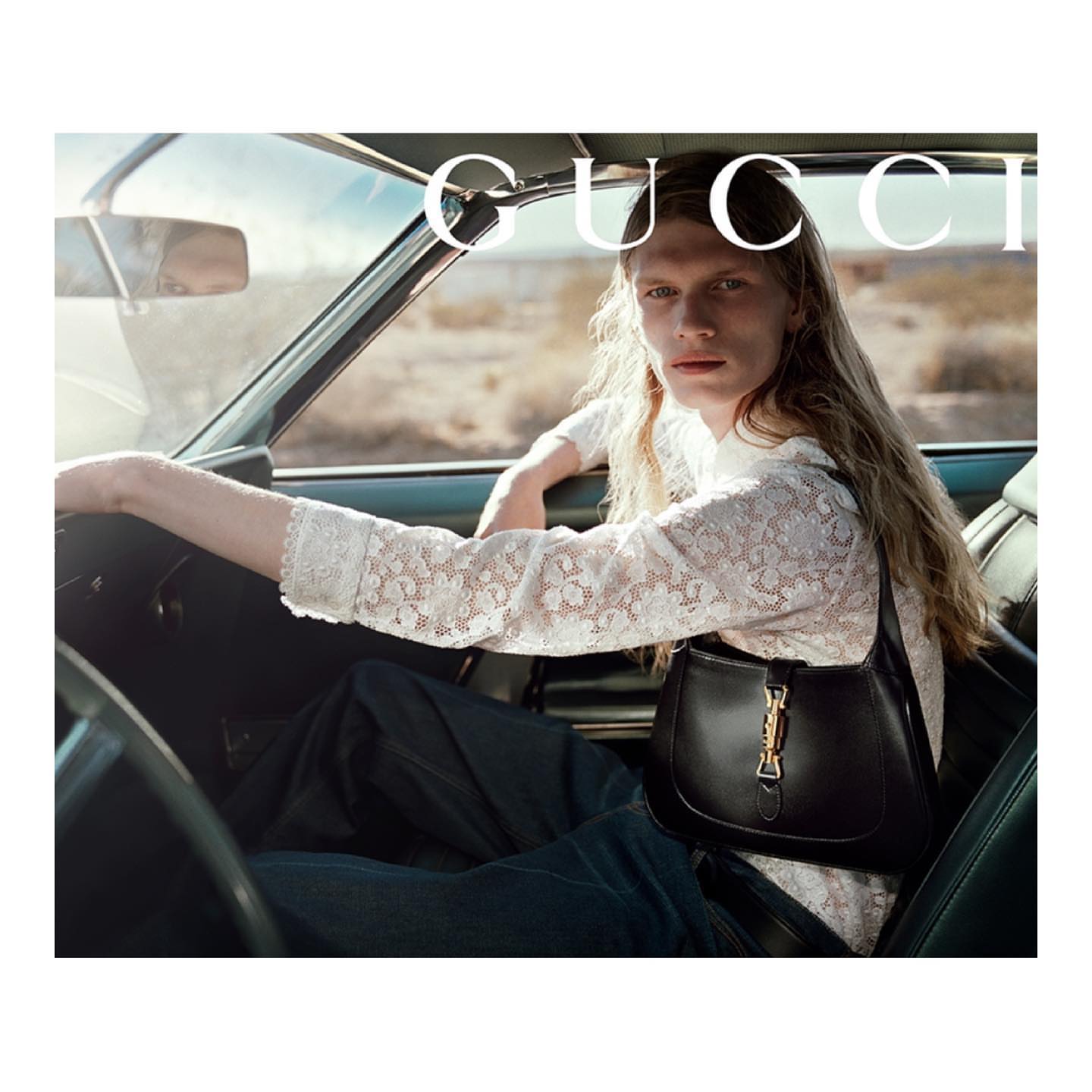 Model with long blonde hair seated inside a car wearing a white lace top and dark pants, holding a black leather handbag.
