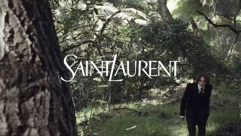 Man in a black suit walking through a dense forest with Saint Laurent text overlaid across the woodland background