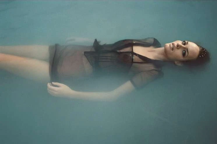 Adult woman floating in tranquil water wearing a sheer black top. Underwater shot with soft, diffused lighting creates a serene atmosphere.