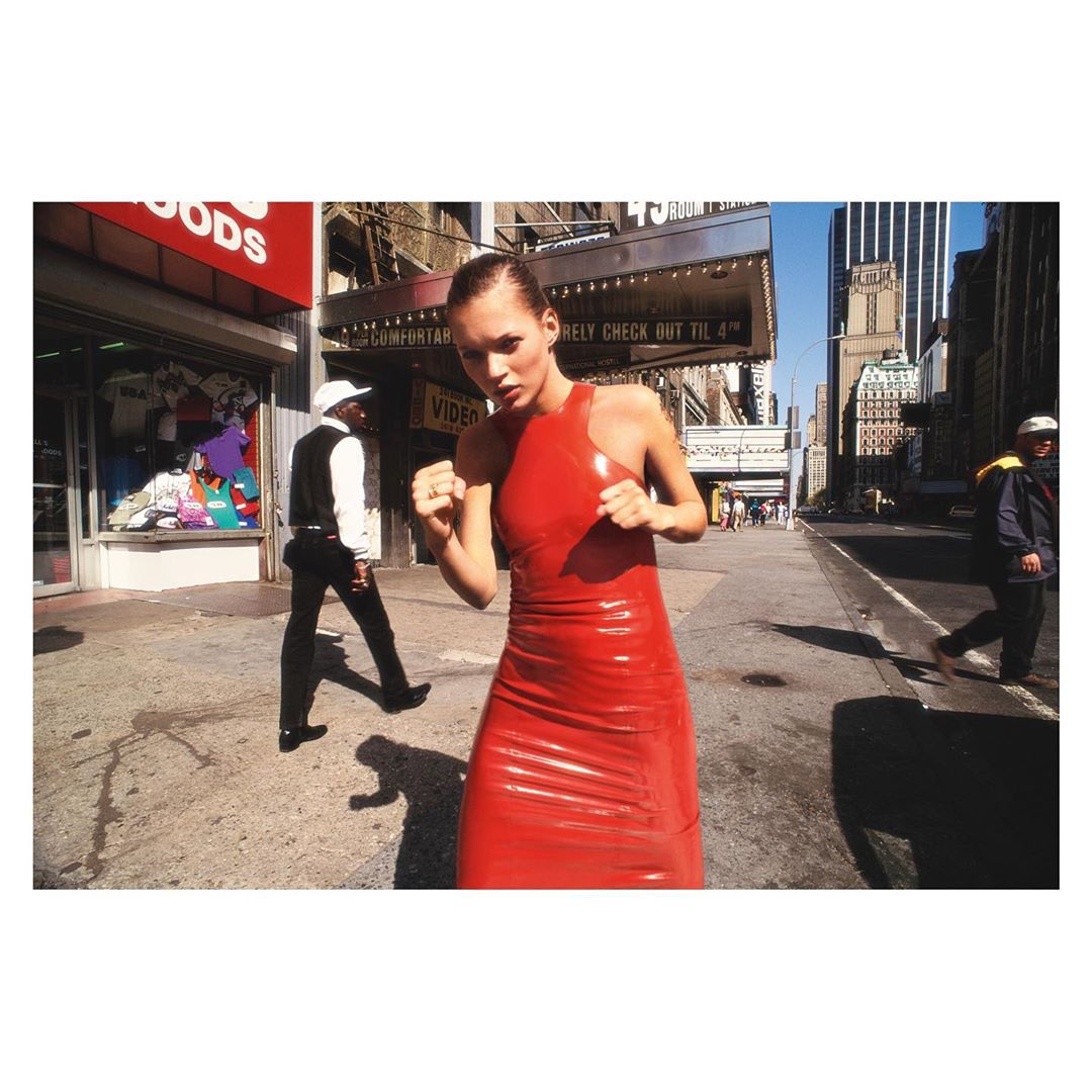 Fashion shot of Kate Moss in a red latex dress posing in a fighting stance on a city street, with pedestrians and buildings in the background.