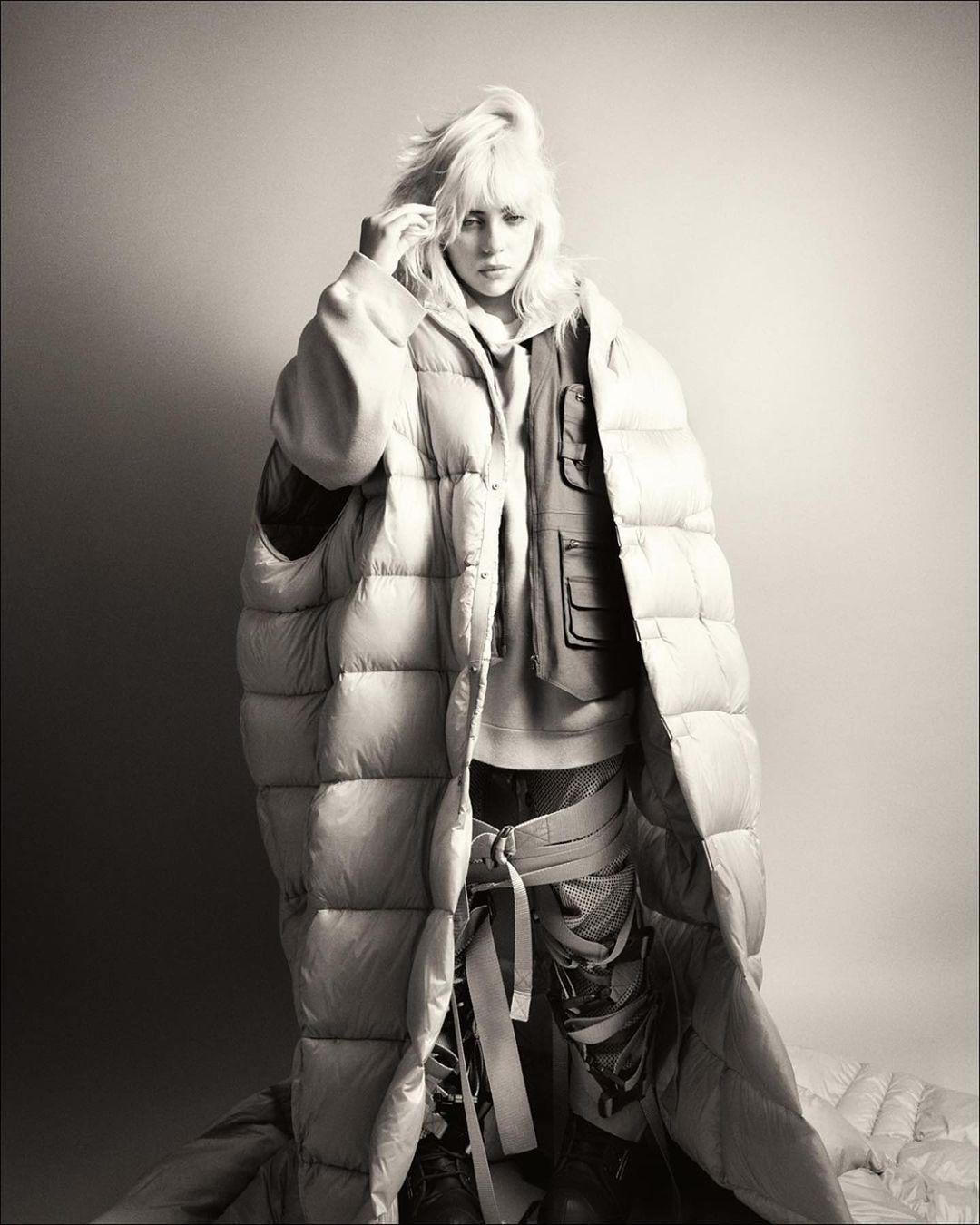 Black-and-white portrait of Billie Eilish in a puffy down jacket with white-blonde hair, posing against a gradient grey background.