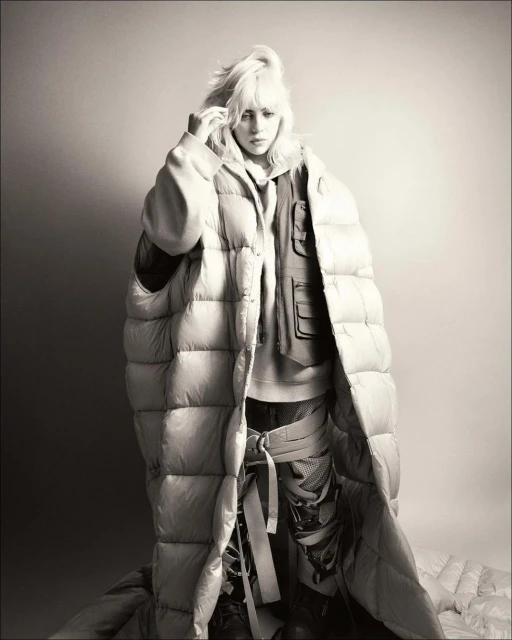 Black-and-white portrait of Billie Eilish in a puffy down jacket with white-blonde hair, posing against a gradient grey background.