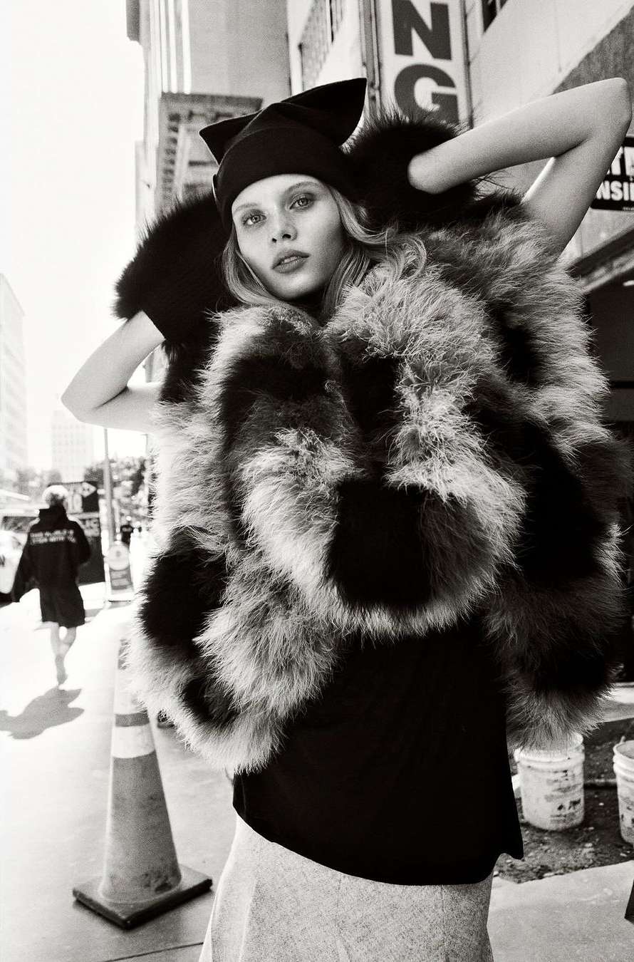 A woman in an oversized fur coat poses outdoors on a city street, wearing a black hat and top, with buildings in the background.