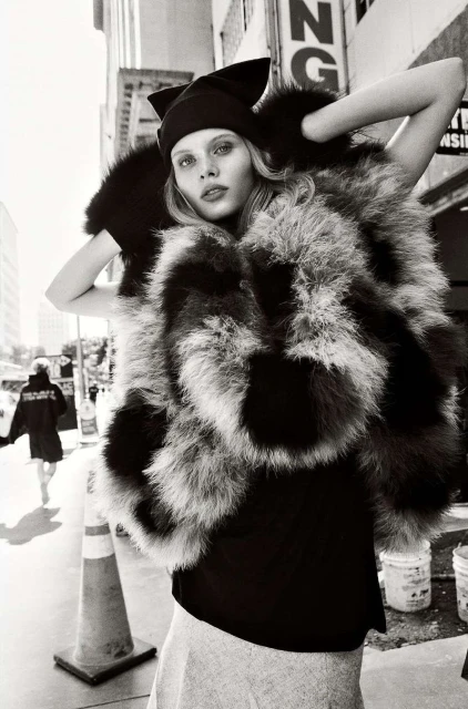 A woman in an oversized fur coat poses outdoors on a city street, wearing a black hat and top, with buildings in the background.