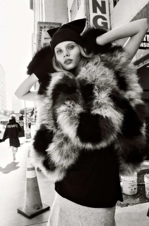 A woman in an oversized fur coat poses outdoors on a city street, wearing a black hat and top, with buildings in the background.