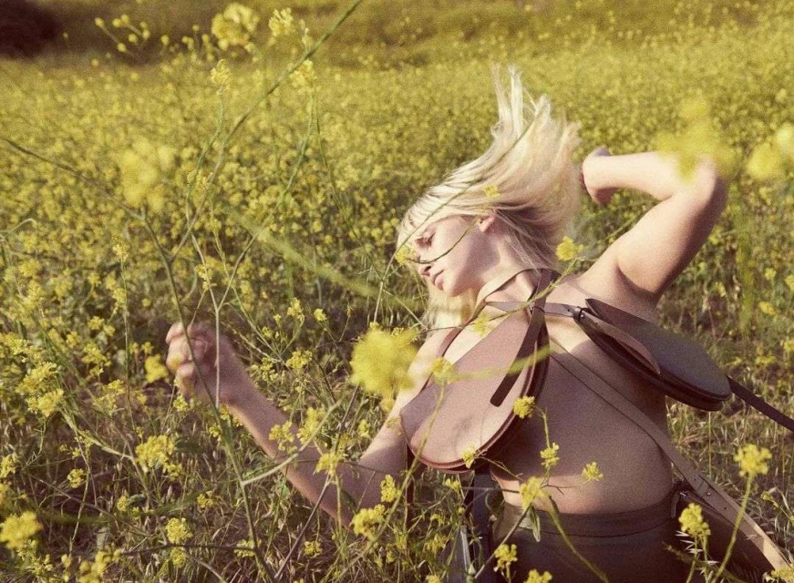 Woman with blonde hair and olive skin poses in a field of yellow flowers wearing a vest and shoulder bag in an outdoor setting.