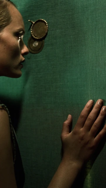 Woman in profile with hand on a green wall and spy-hole-style decoration, low-key lighting and desaturated colors, moody and mysterious.