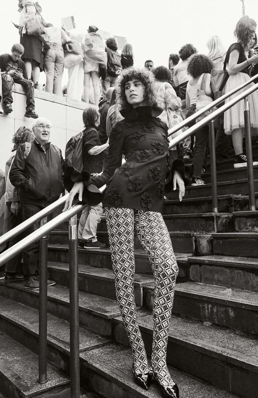 Model in patterned leggings and dark blouse posing on outdoor stone stairs surrounded by a crowd.
