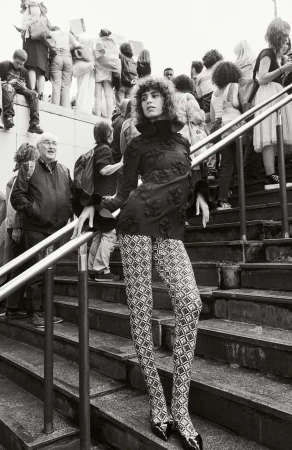 Model in patterned leggings and dark blouse posing on outdoor stone stairs surrounded by a crowd.
