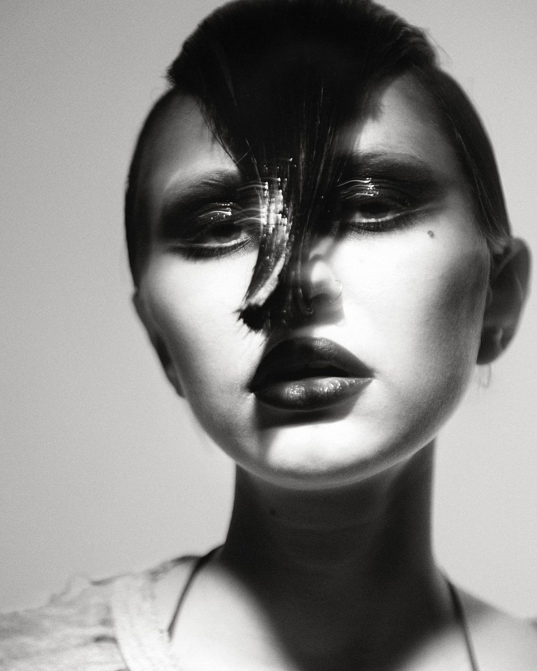 Monochrome close-up portrait of a woman with stylized dark hair and bold makeup against a neutral background in a photo studio.