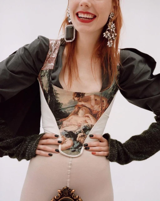 A smiling woman with auburn hair wears a vintage-inspired top with art images, large earrings, and black sleeves against a white backdrop.