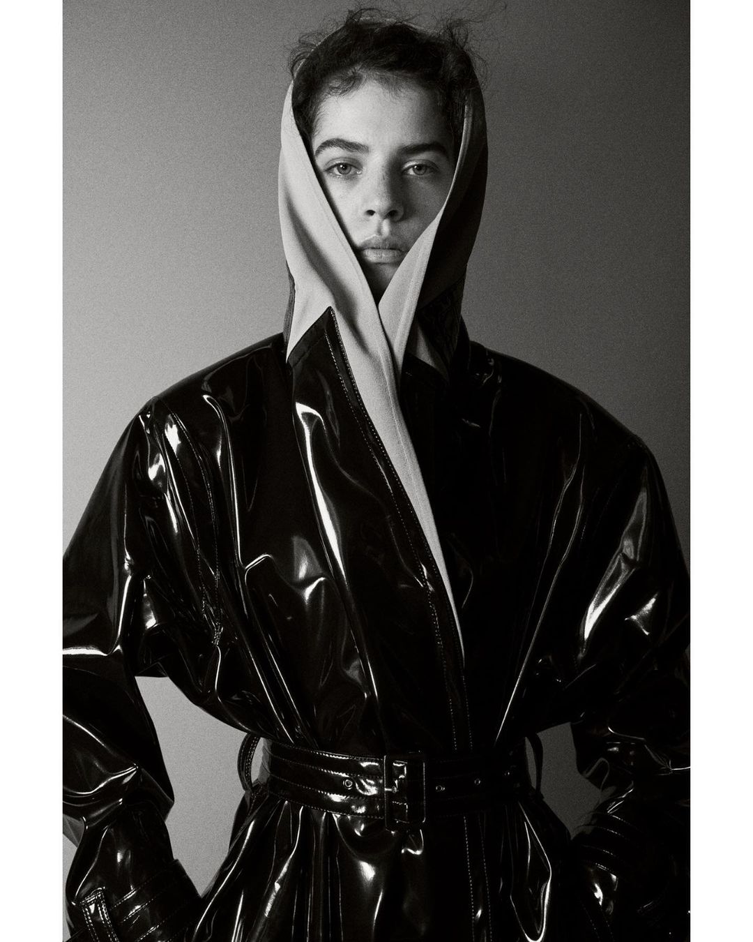 Monochrome portrait of a person with a neutral facial expression, wearing a black patent leather trench coat and beige head covering.