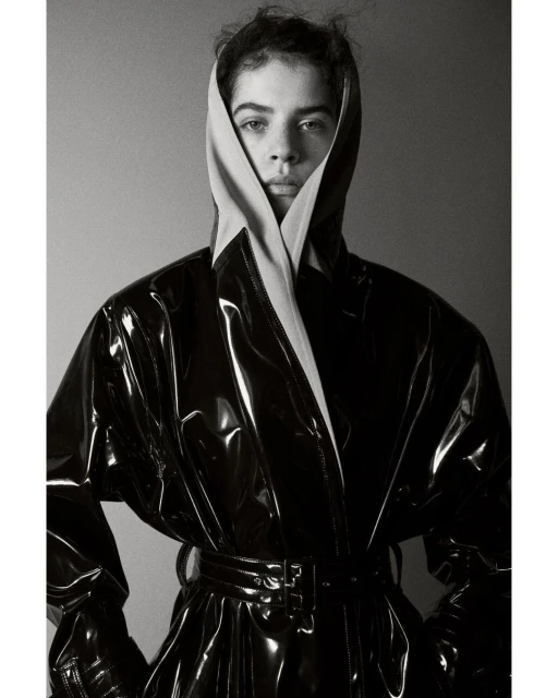 Monochrome portrait of a person with a neutral facial expression, wearing a black patent leather trench coat and beige head covering.