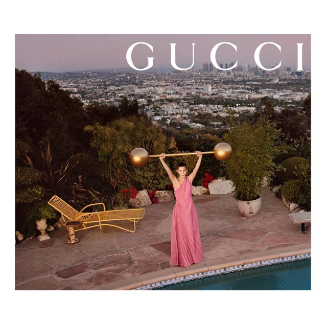 Woman in pink dress poses with gold barbell overlooking Los Angeles skyline. Fashion shot with vintage tones; Gucci branding.
