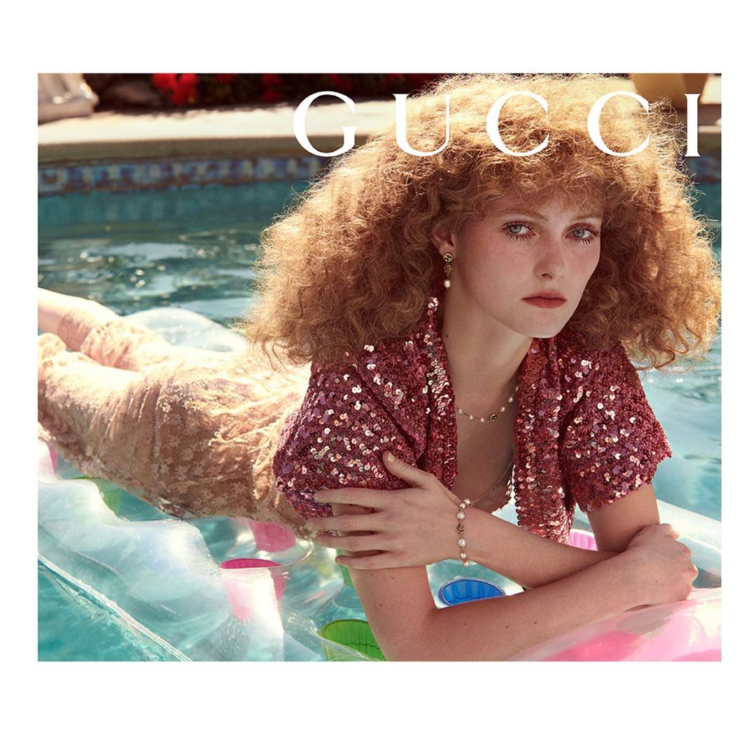 Adult female model with auburn curly hair reclines on an inflatable raft in a swimming pool, wearing a sequined jacket and pearl jewelry.